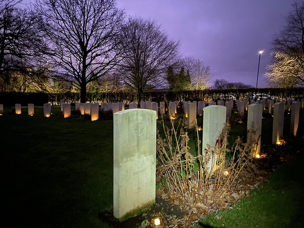 CWGC headstones illuminated from below using temporary lights. The sky is a deep purple. Leafless trees are visible in the background.